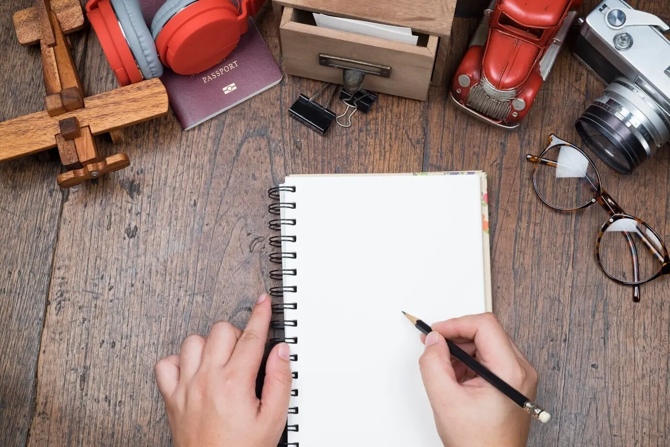 Eine Hand hält einen Bleistift und ein weißes Notizbuch mit leerem Papier auf einem Holztisch. Konzept der Reisevorbereitung.